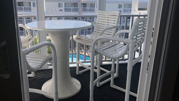 A balcony with a white table and chairs, overlooking a swimming pool and high-rise buildings.
