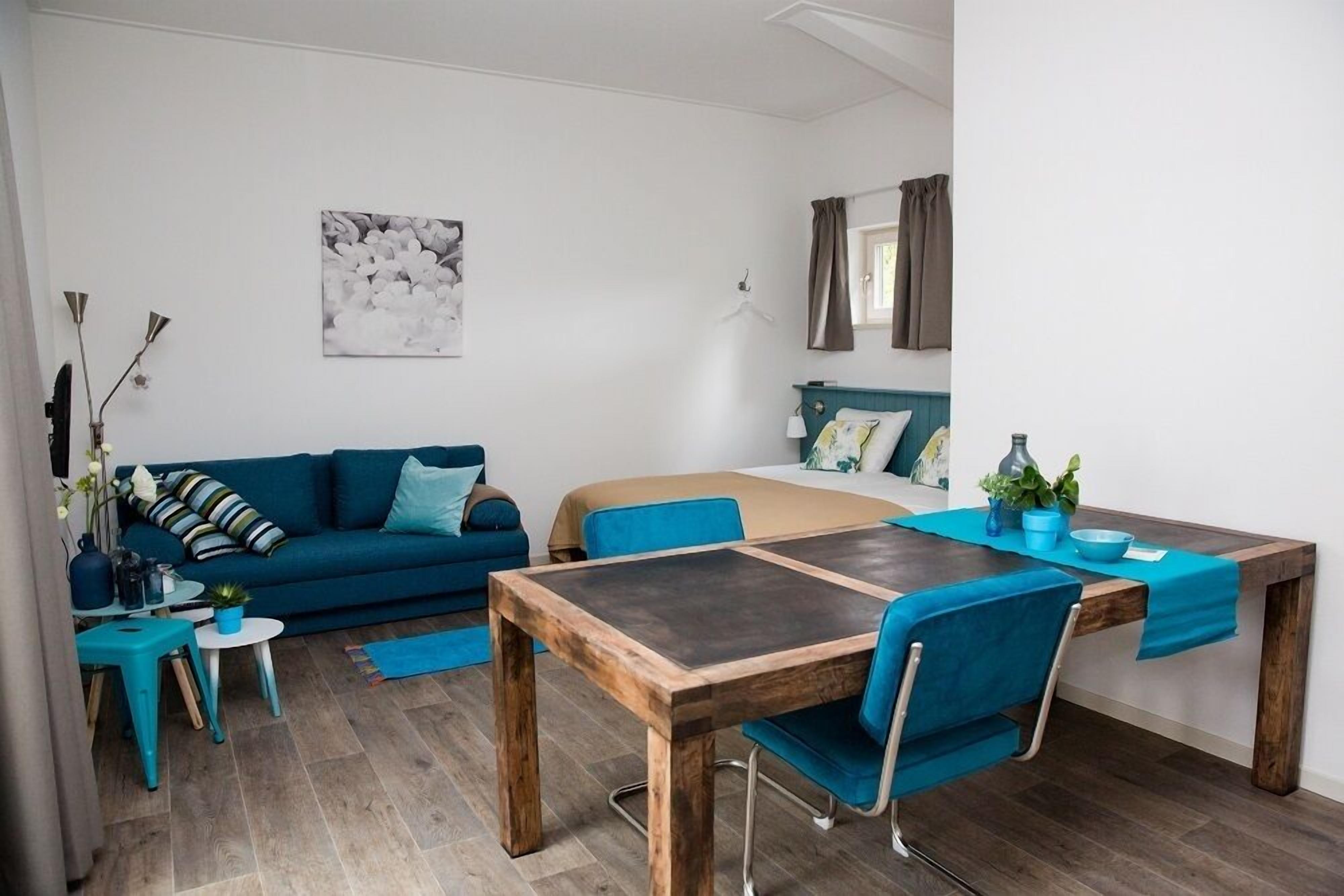 A modern, well-lit room with a blue sofa, a wooden dining table with blue chairs, a bed with a blue headboard, and a small side table with a plant.