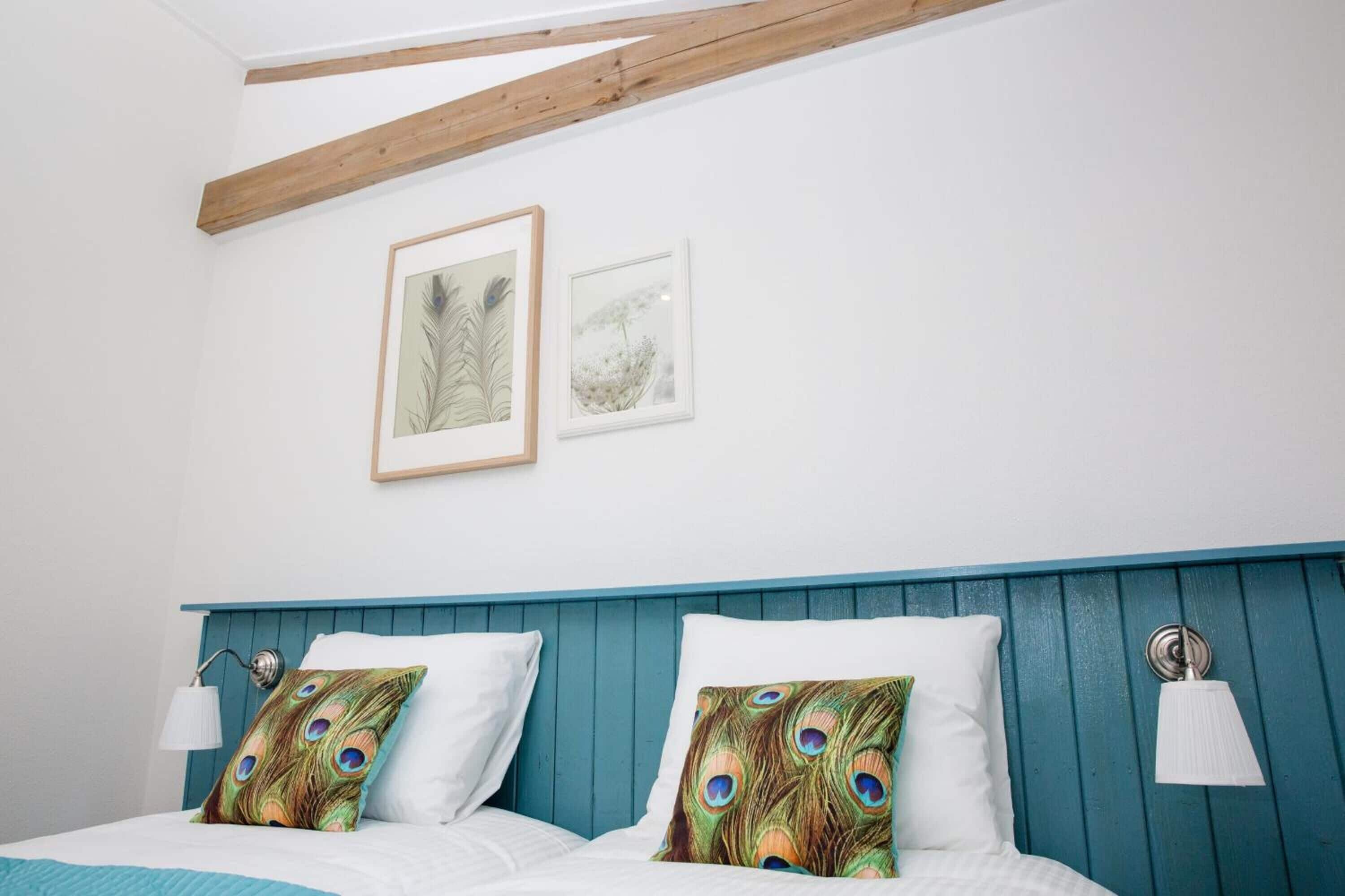 A bedroom with a wooden ceiling, two framed artworks, a bed with white bedding, and peacock-themed pillows.