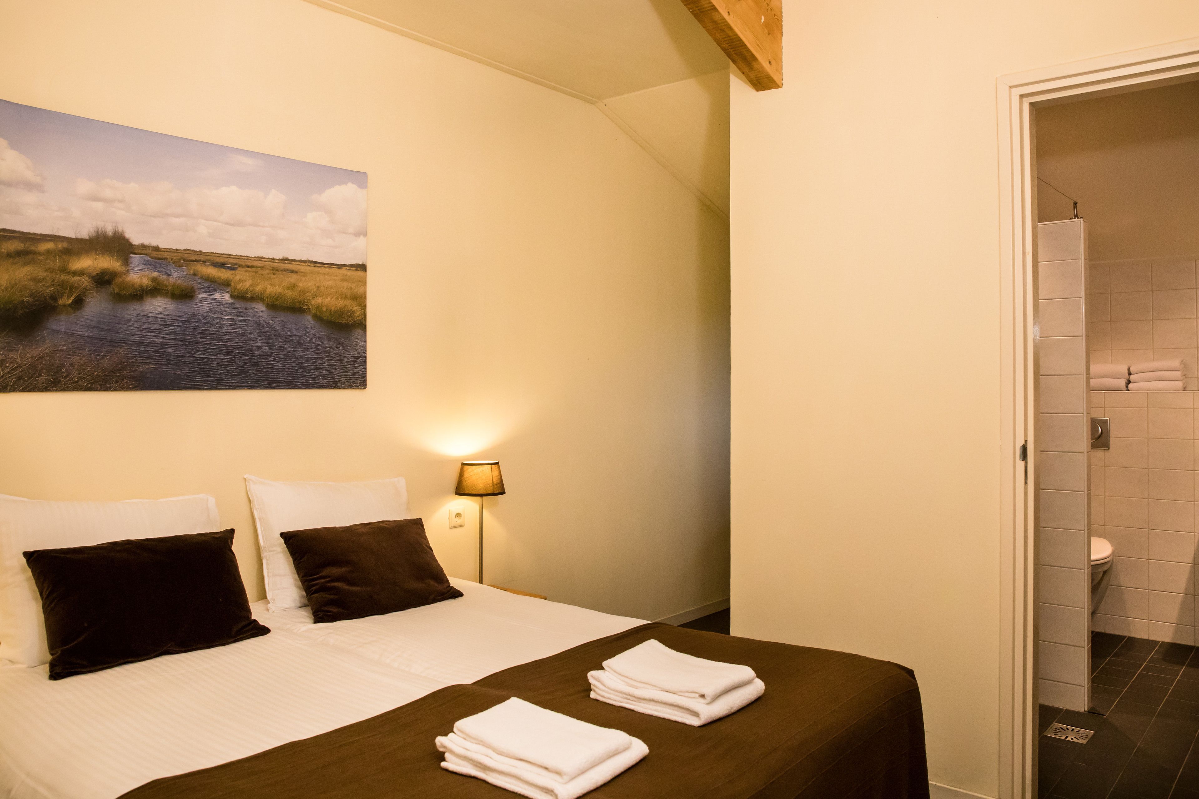A hotel room with a bed, pillows, a painting of a landscape, a lamp, and a bathroom visible through an open door.
