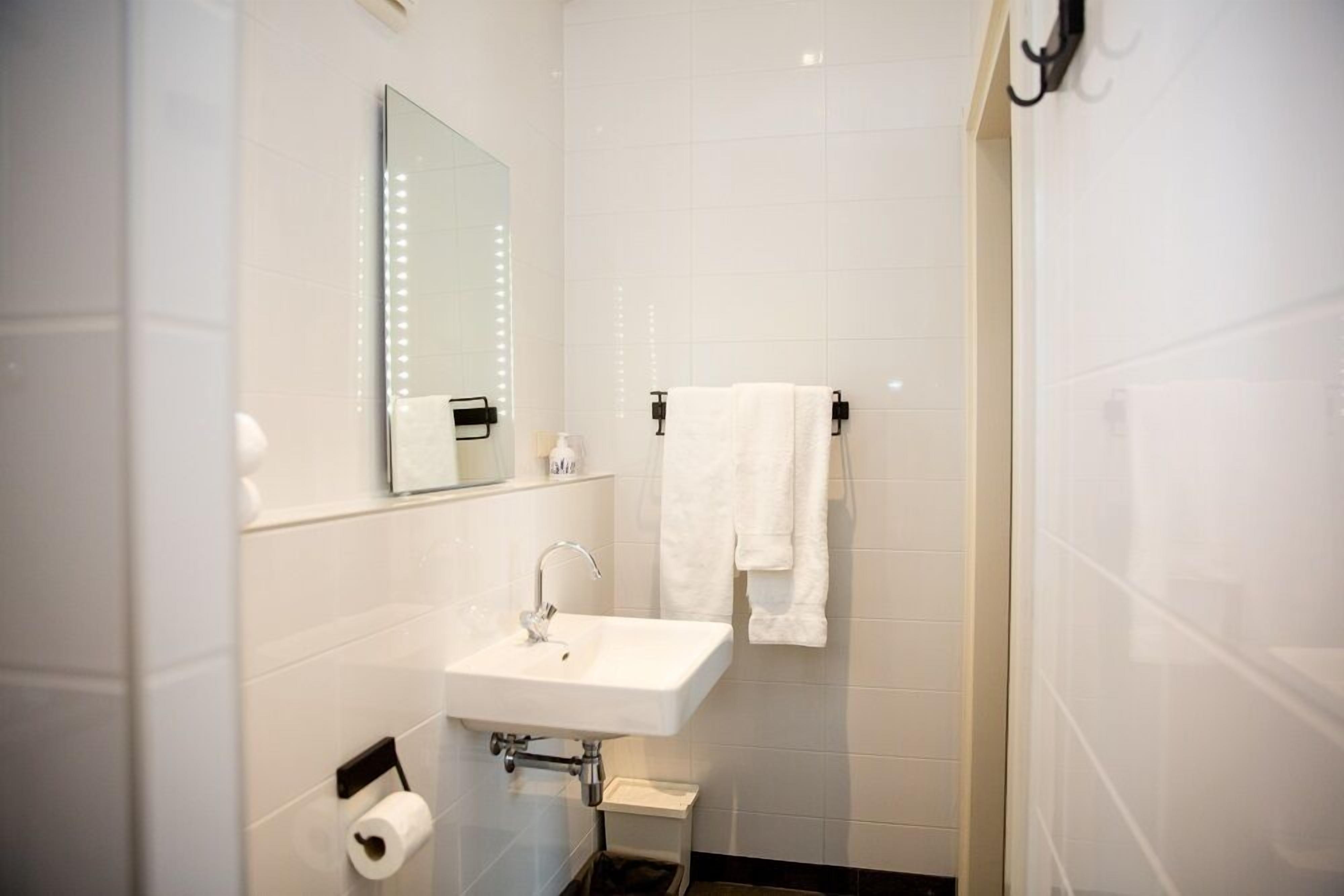 A modern bathroom with a white sink, a mirror with lights, a towel rack, and a toilet paper holder.