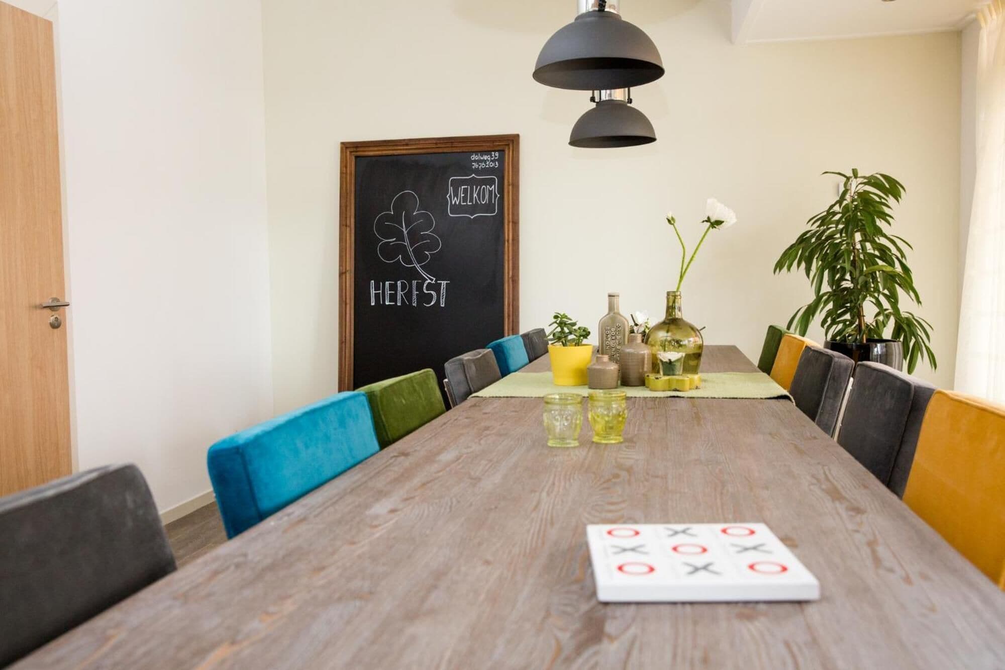 A dining area with a long wooden table, black and yellow chairs, and a chalkboard with 'WELCOME HERST' written on it.