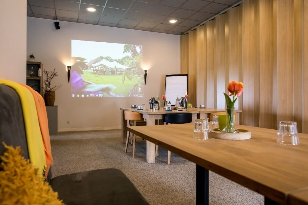 Un salón de conferencias con una mesa de madera, sillas, una pantalla de proyector y un jarrón con flores.