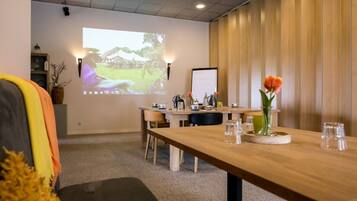 Un salón de conferencias con una mesa de madera, sillas, una pantalla de proyector y un jarrón con flores.