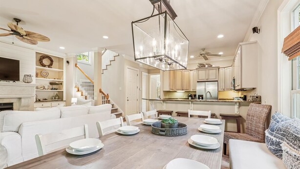A spacious dining area with a wooden table set for six, surrounded by white chairs. A modern chandelier hangs above, and a staircase with wooden railings leads to an upper level.