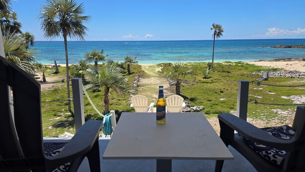 Una mesa con sillas frente a una vista de la playa, una botella de cerveza Kalik y una falda hula.