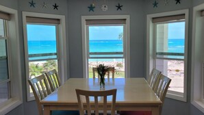 Una mesa de comedor con cuatro sillas, un florero con flores y vista a la playa a través de amplias ventanas.