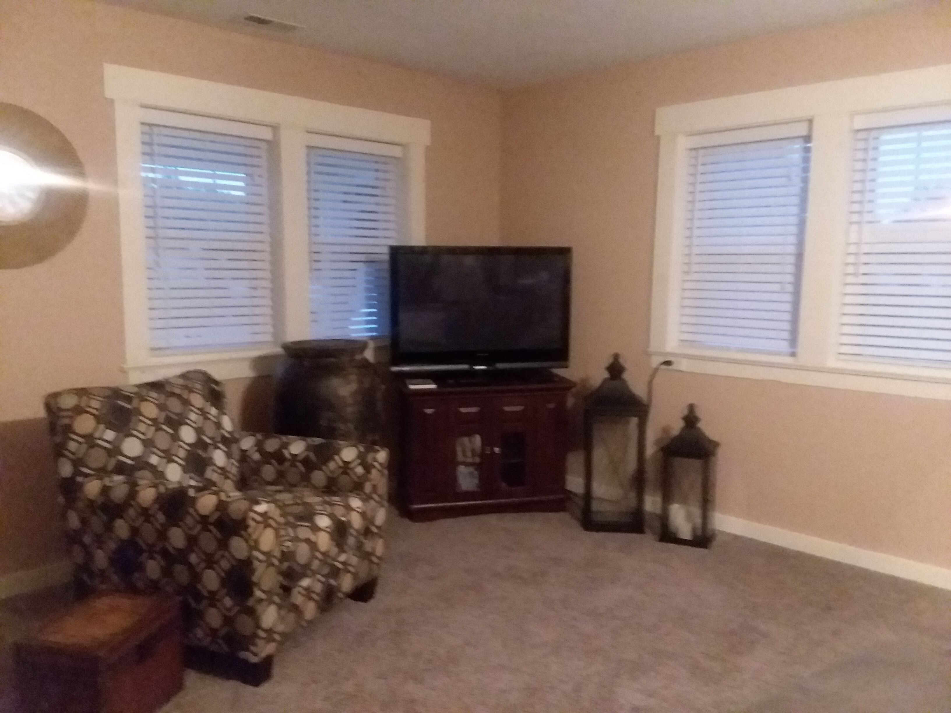 Una sala de estar con un sofá, un televisor sobre un mueble bajo, dos ventanas con persianas y dos lámparas de pie.