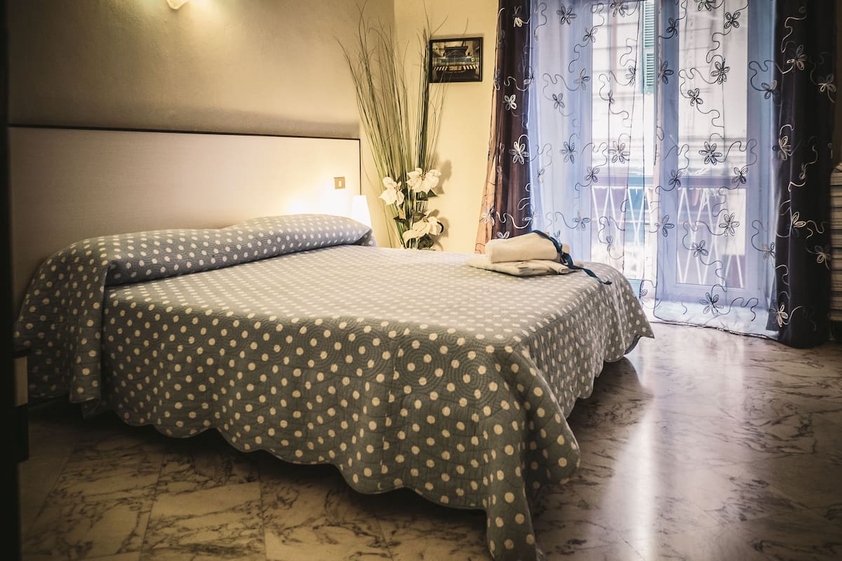 Une chambre d’hôtel avec un grand lit, une table de chevet, un vase de fleurs et une vue sur l’extérieur à travers des rideaux transparents.