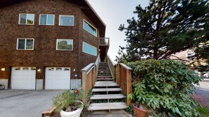 A two-story wooden building with a staircase leading to a balcony. There are two garage doors and a potted plant with a red flower at the base of the stairs.