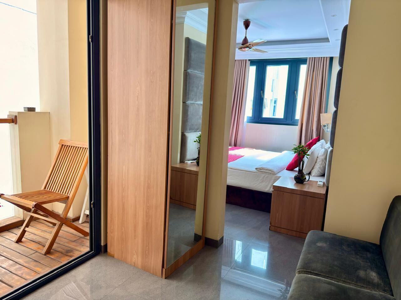 Hotellihuone, jossa on puinen tuoli, sängyn vieressä yöpöytä, ikkunassa verhot ja katossa kattotuuletin.