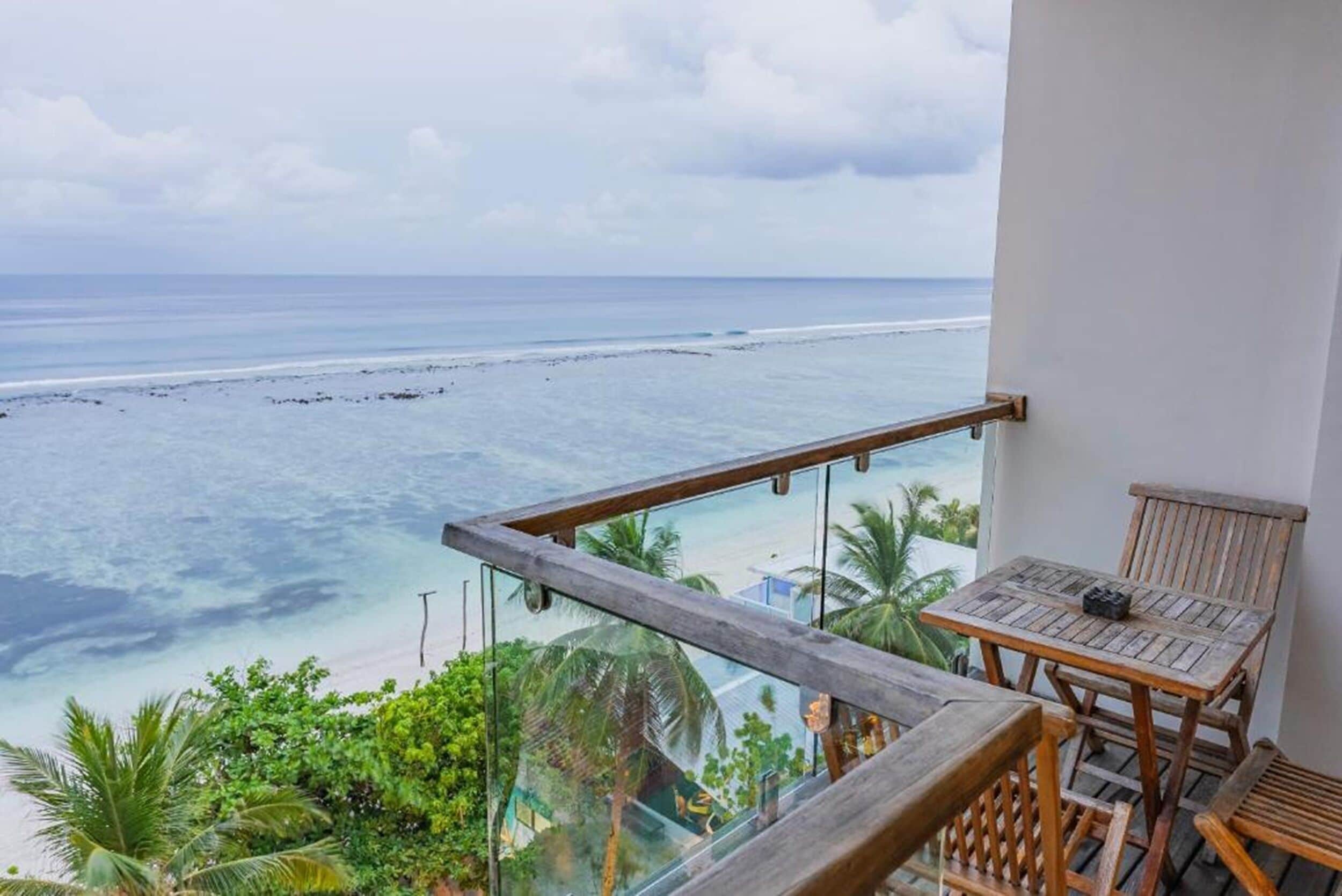 A balcony with a view of the ocean and beach, wooden furniture, and tropical plants.