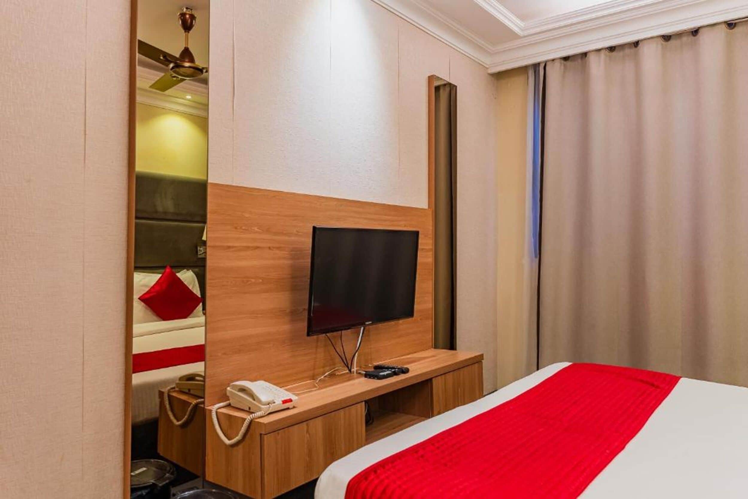 Habitación de hotel con cama, televisor montado en un panel de madera, teléfono en la mesita de noche y una pequeña mesa con lámpara.