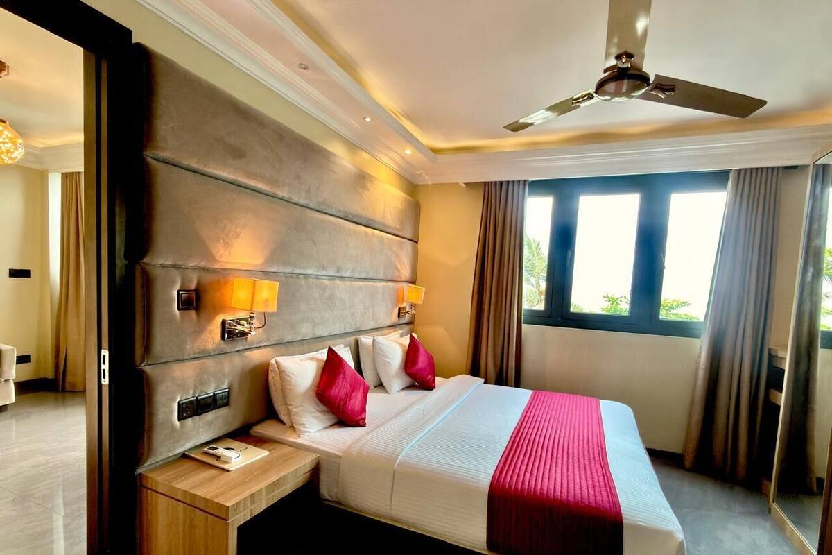 Quarto de hotel com uma cama grande, luminárias de cabeceira, mesa de cabeceira, ventilador de teto e janela com cortinas.