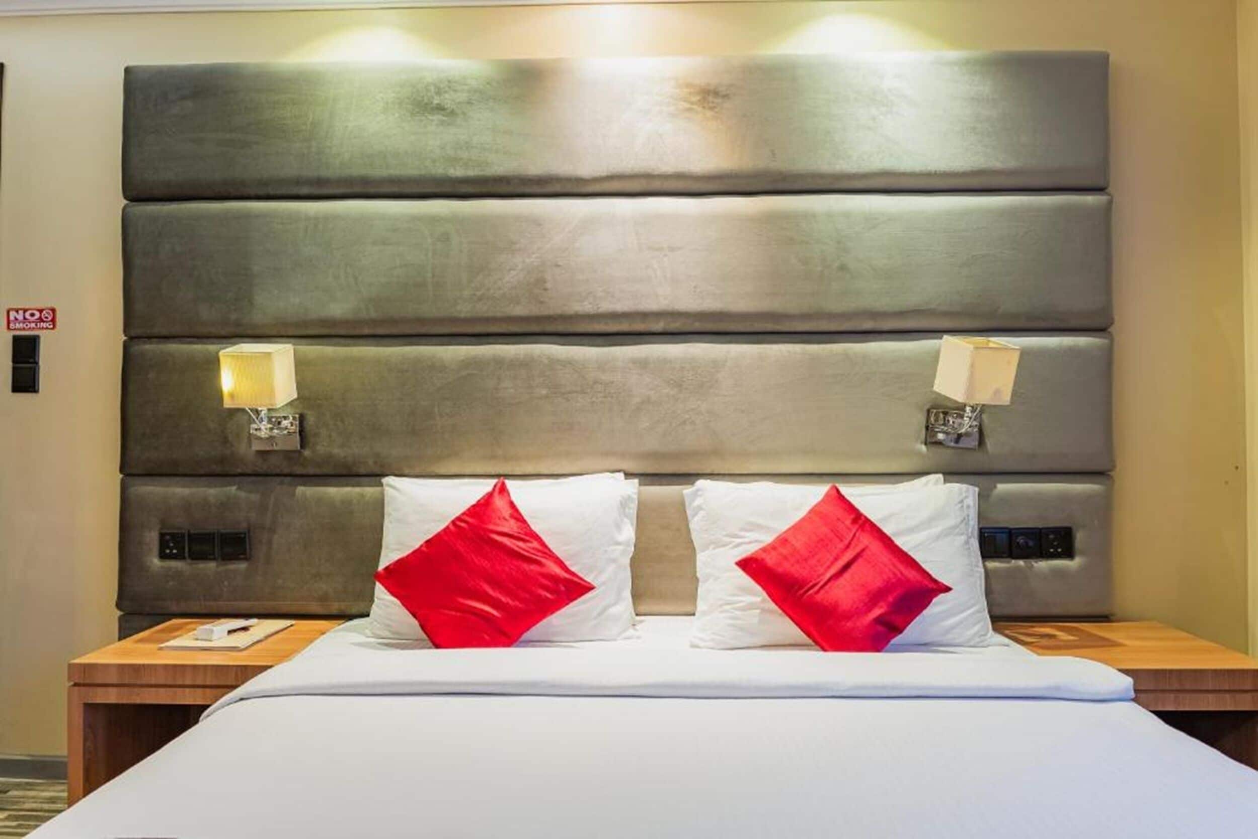 Een hotelkamer met een groot, bekleed hoofdeinde, twee bedlampen, een bed met witte beddengoed en rode kussens, en een nachtkastje van hout met een boek.
