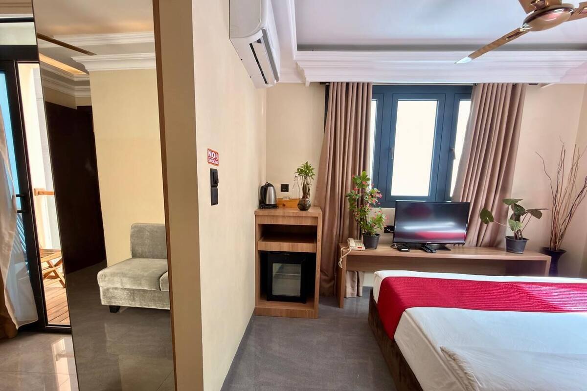 Quarto de hotel com cama, escrivaninha com televisão, poltrona, lareira pequena e janela com cortinas.