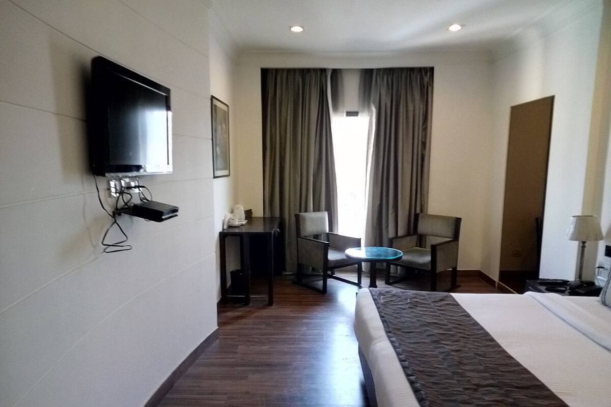Habitación de hotel con cama, televisor montado en la pared, escritorio con silla y ventana con cortinas.