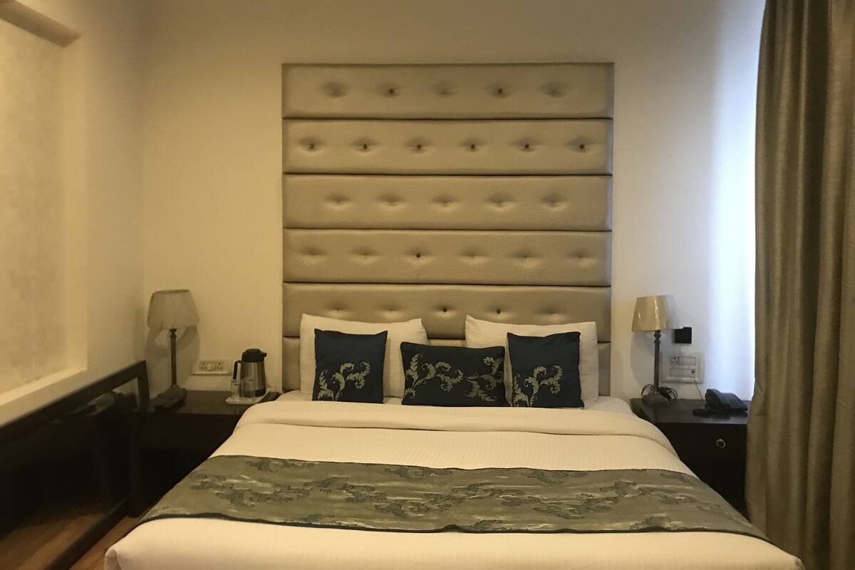 Habitación de hotel con una cama grande, cabecera con botones, dos mesitas de noche con lámparas y una ventana con cortinas.