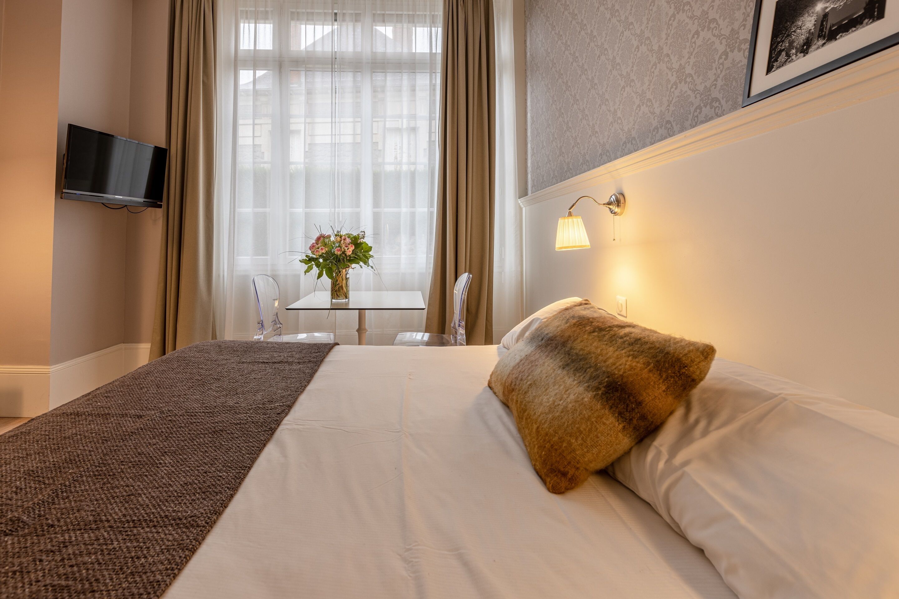 Een hotelkamer met een bed, een aan de muur bevestigde televisie, een raam met gordijnen, een tafeltje met een bloevas en een wandlamp.