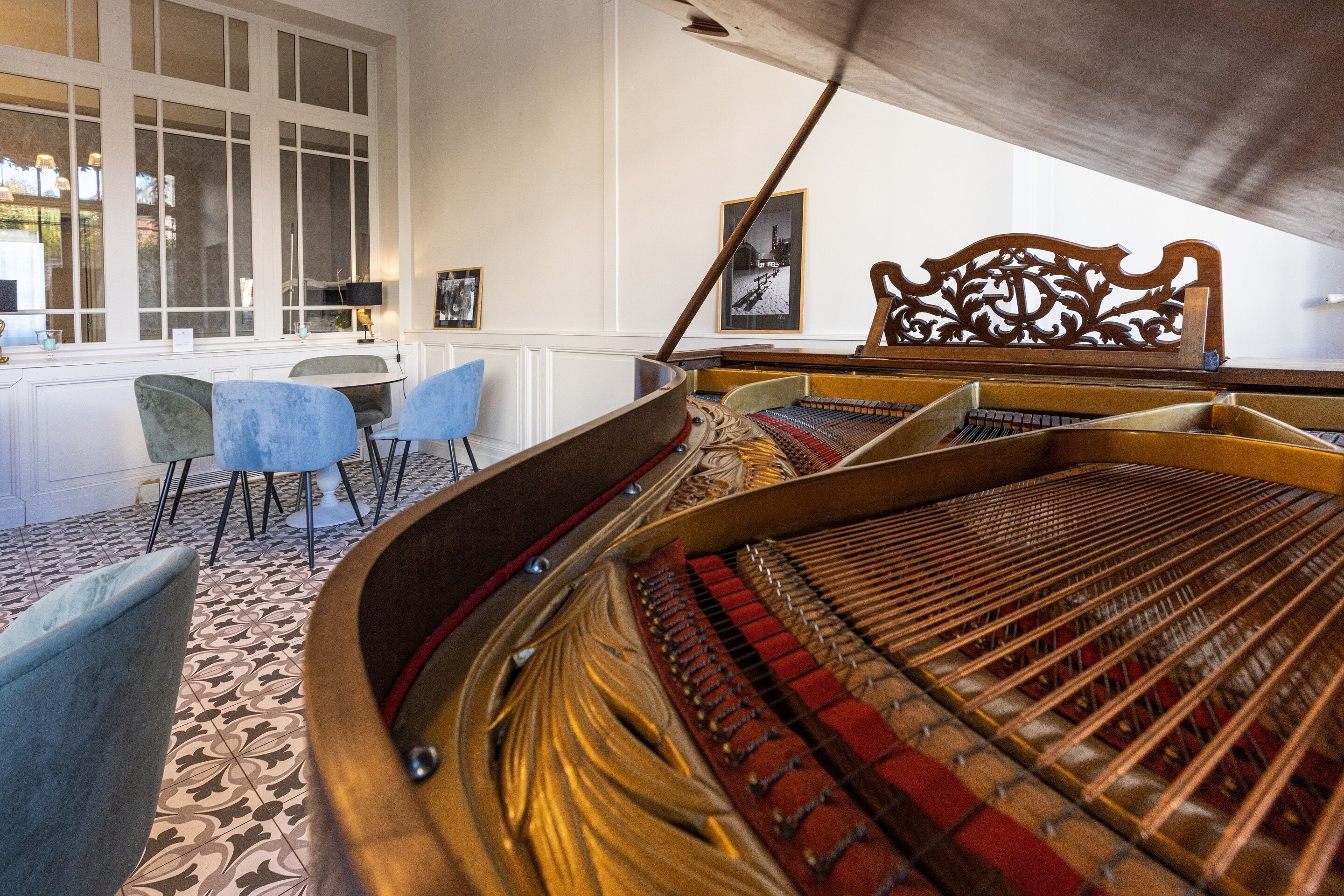 A grand piano in a room with patterned carpet, a wooden chair with intricate backrest, and a table with chairs.