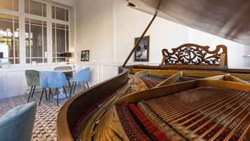Un piano à queue dans une pièce dont le sol est recouvert d’un tapis à motifs, une chaise en bois au dossier finement travaillé et une table entourée de chaises.