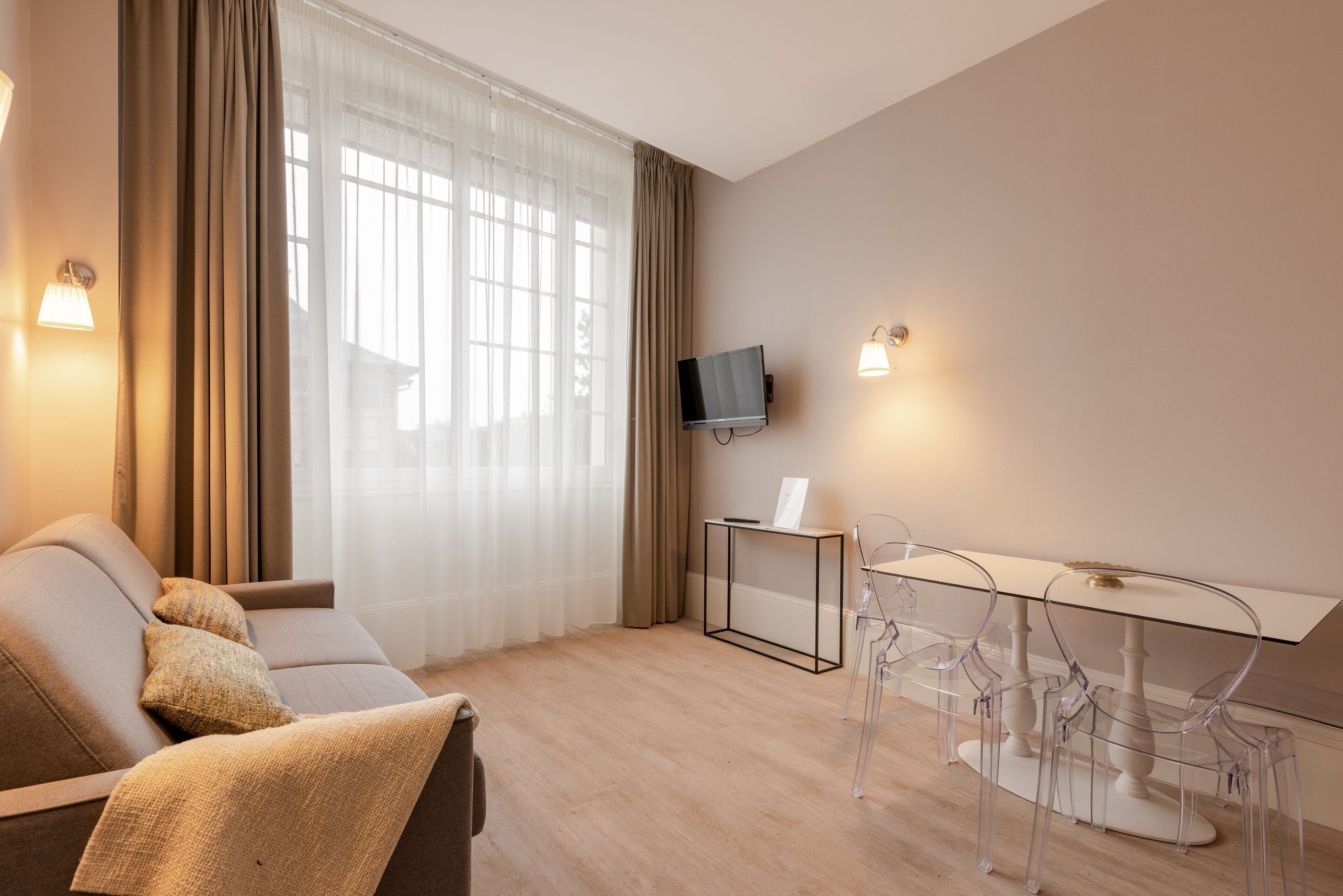 Habitación de hotel moderna con sofá, una mesa de comedor de vidrio, televisor montado en la pared y una ventana con cortinas translúcidas.