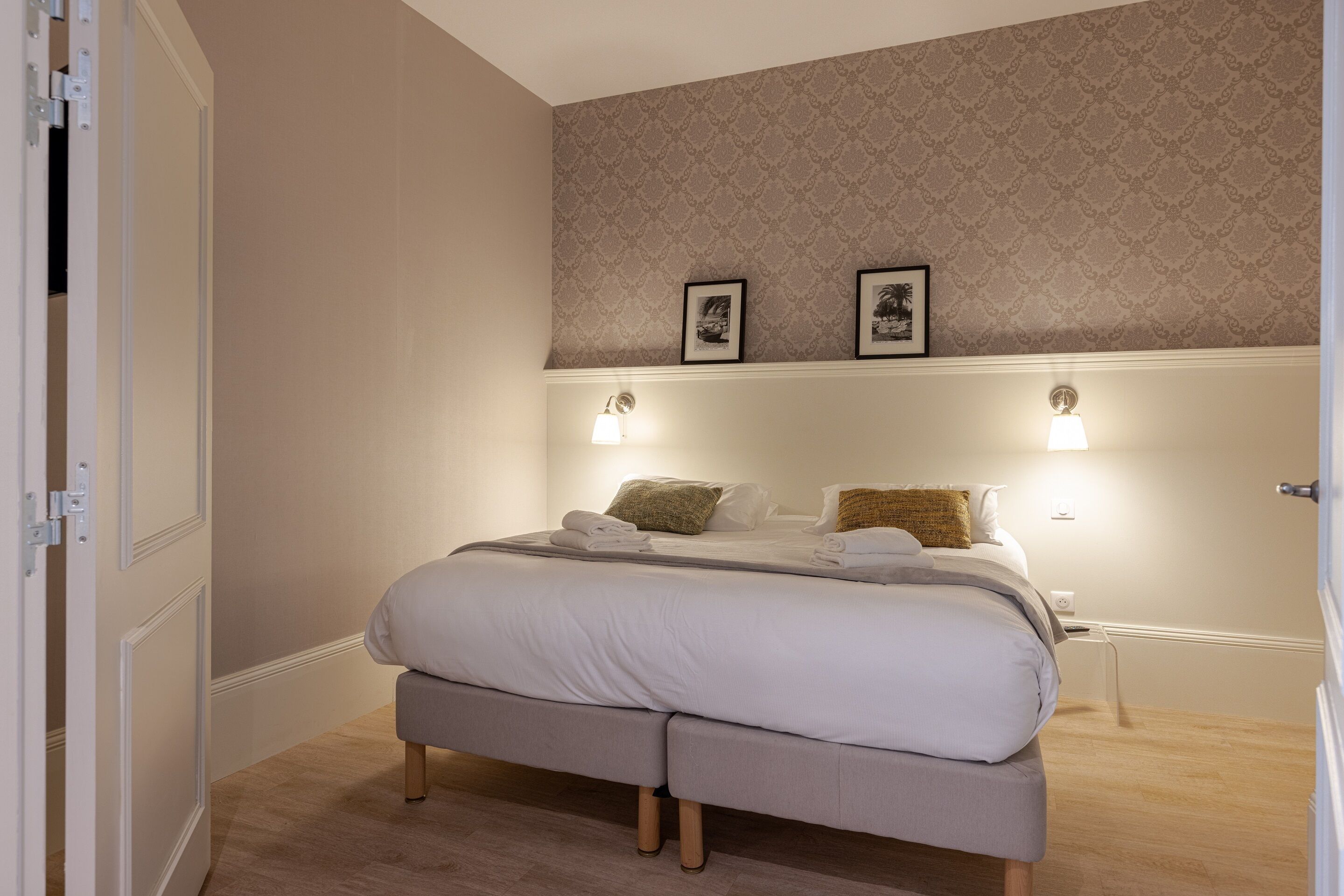 Una cama bien hecha con sábanas blancas y dos almohadas, una mesita de noche con una lámpara y un papel tapiz estampado en la pared.