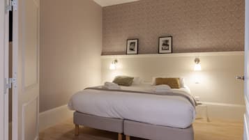 Una cama bien hecha con sábanas blancas y dos almohadas, una mesita de noche con una lámpara y un papel tapiz estampado en la pared.