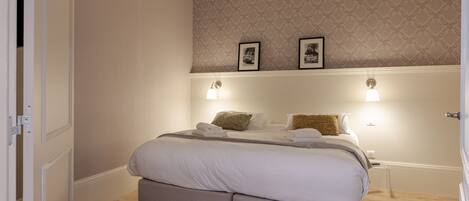 A neatly made bed with white linens and two pillows, a bedside table with a lamp, and a patterned wallpaper on the wall.