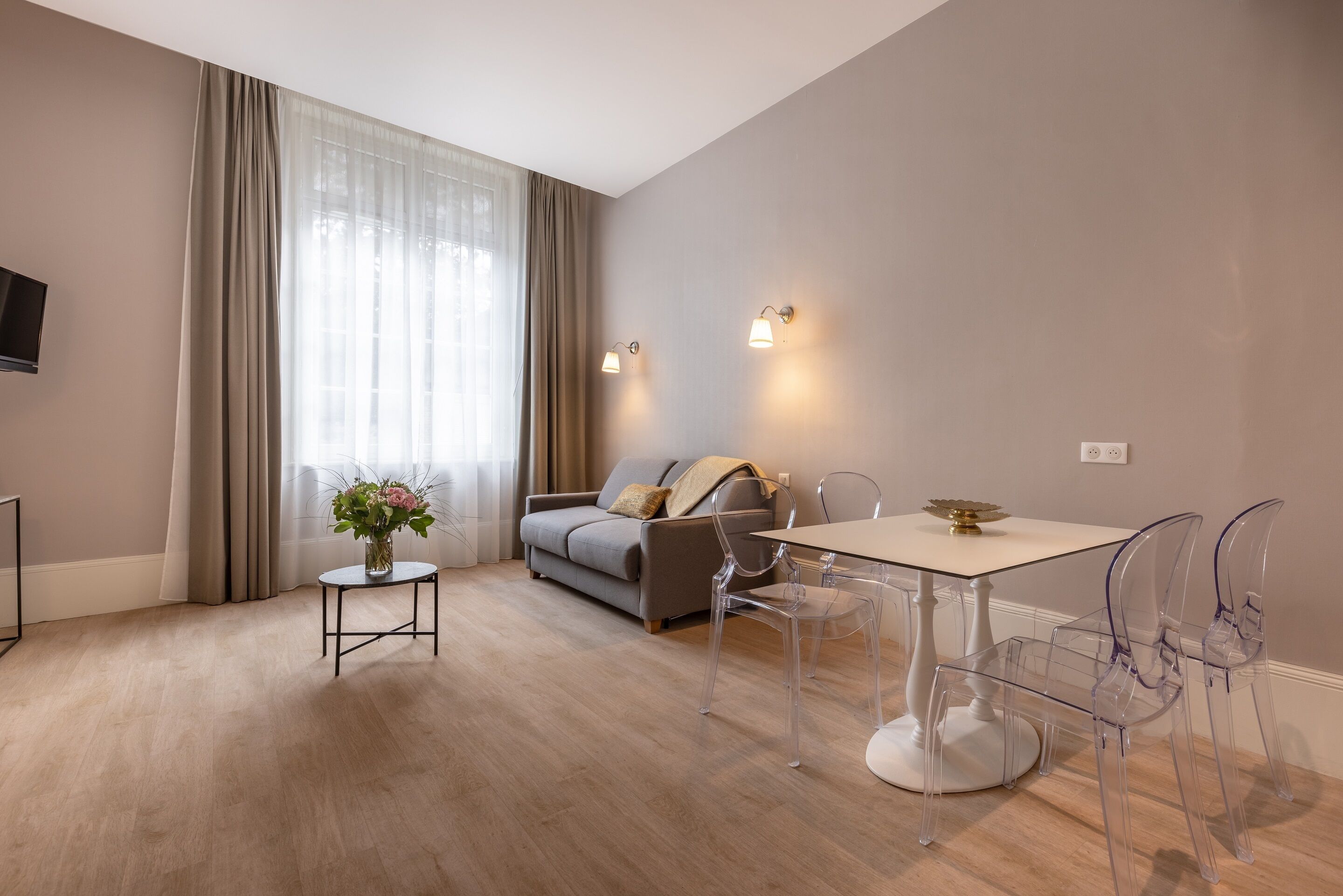 A modern living room with a sofa, a dining table with chairs, a small side table with a vase, and a wall-mounted light fixture.