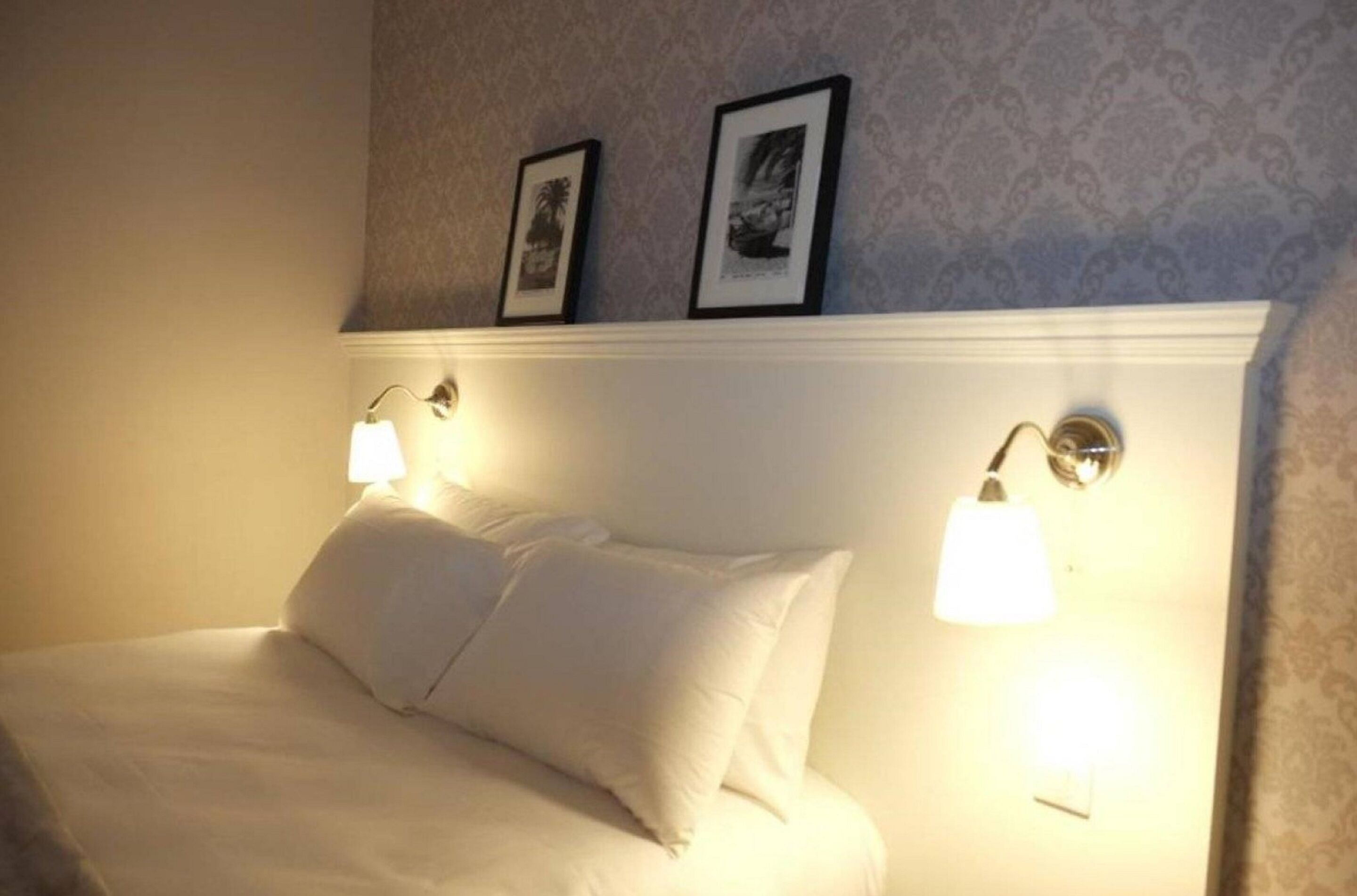 Una cama bien tendida con sábanas blancas y dos lámparas fijadas a la pared a cada lado.