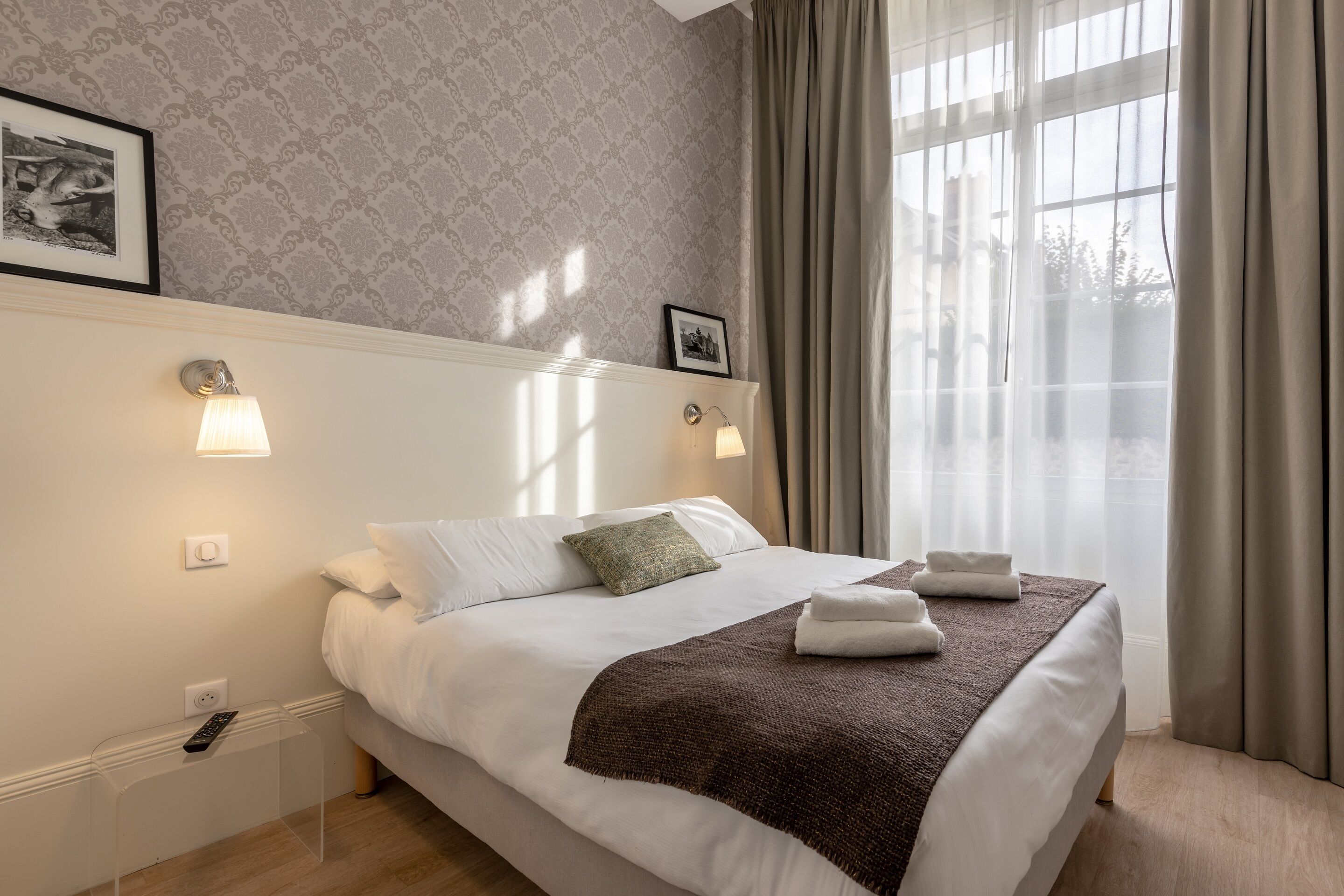A neatly made bed with white linens and a brown throw, a bedside table with a remote control, and a window with sheer curtains.