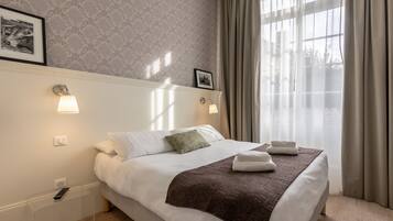 A neatly made bed with white linens and a brown throw, a bedside table with a remote control, and a window with sheer curtains.