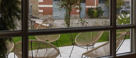 A view from inside a building looking out to a patio area with wicker chairs and a brick building in the background.