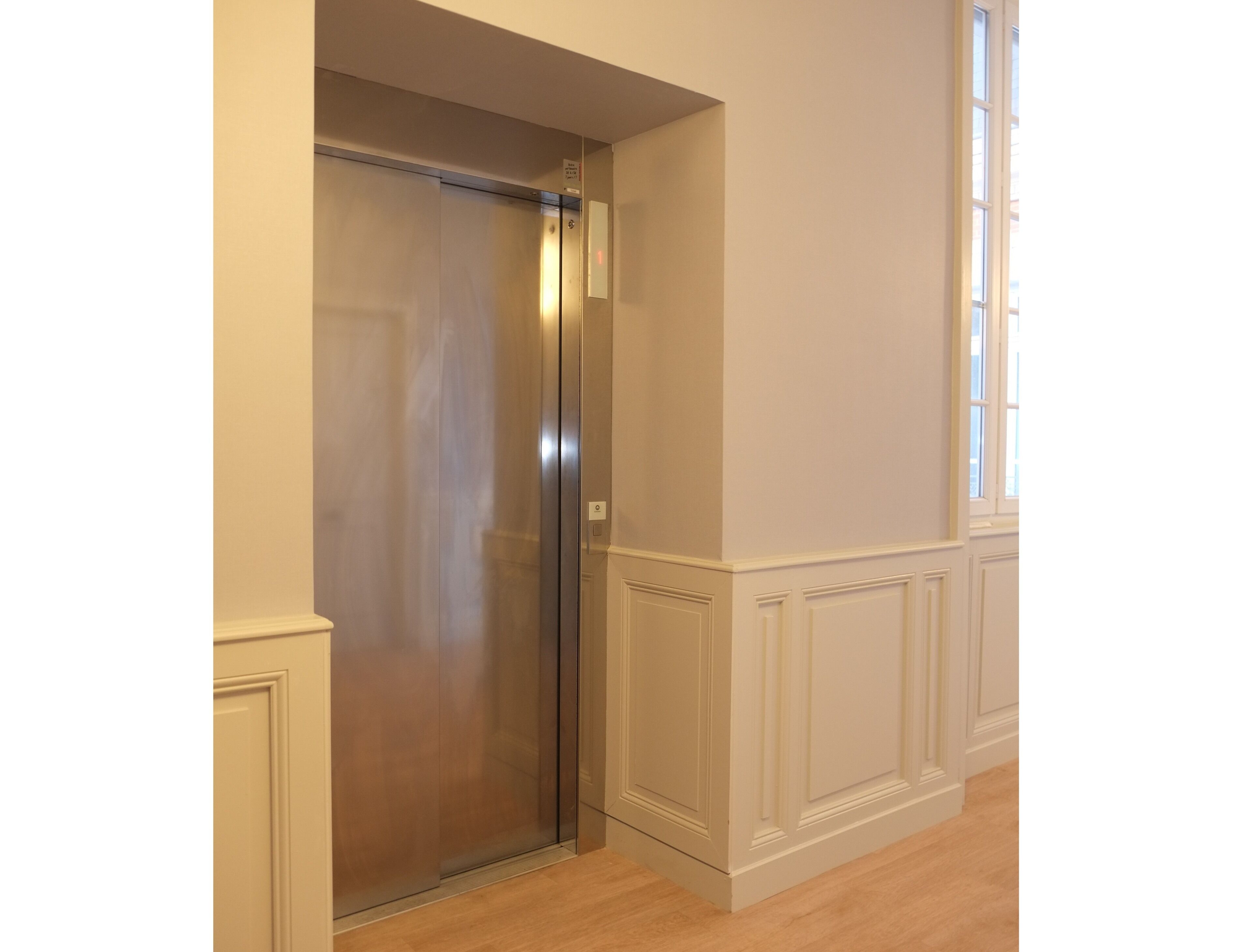 Une porte de monte-charge fermée dans un couloir aux murs recouverts de lambris blancs, avec une fenêtre sur le côté droit.