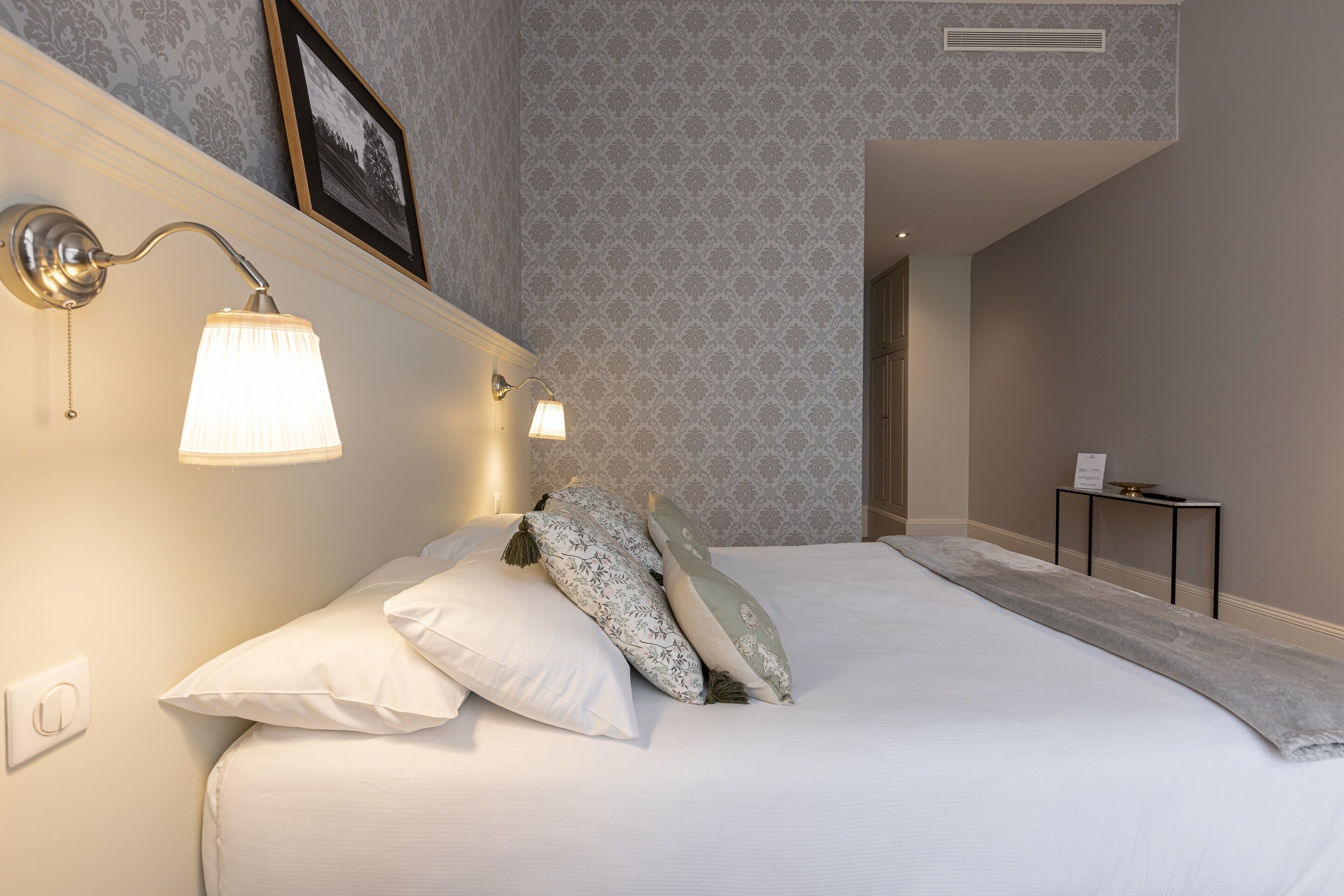 A neatly made bed with white linens and decorative pillows, a wall-mounted lamp, and a small table with a tray.