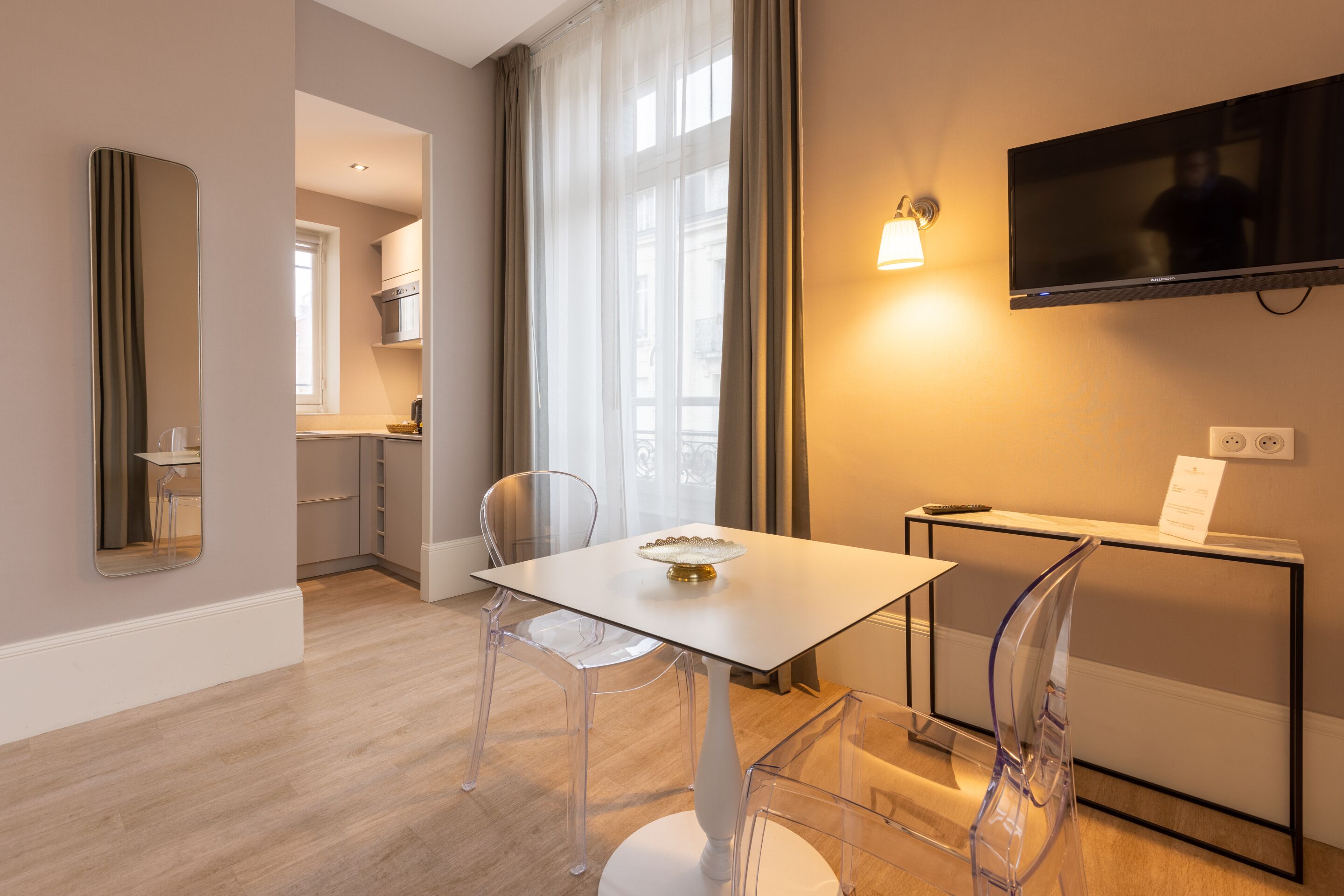 A modern hotel room with a dining area, a TV mounted on the wall, and a view of the city.
