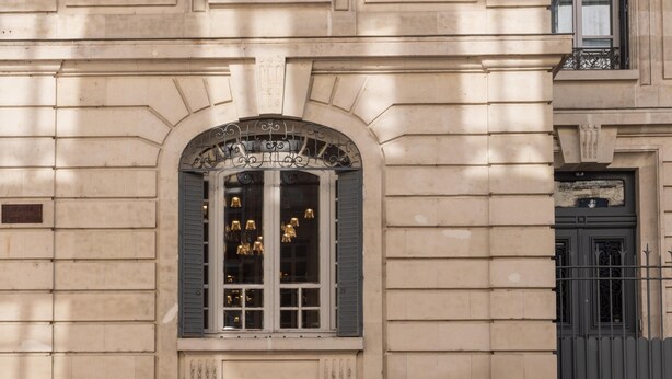 Une façade d’immeuble ornée d’une grande fenêtre en arc surmontée de plusieurs luminaires, d’un balcon doté d’une balustrade en fer forgé et d’une porte agrémentée de motifs métalliques décoratifs.