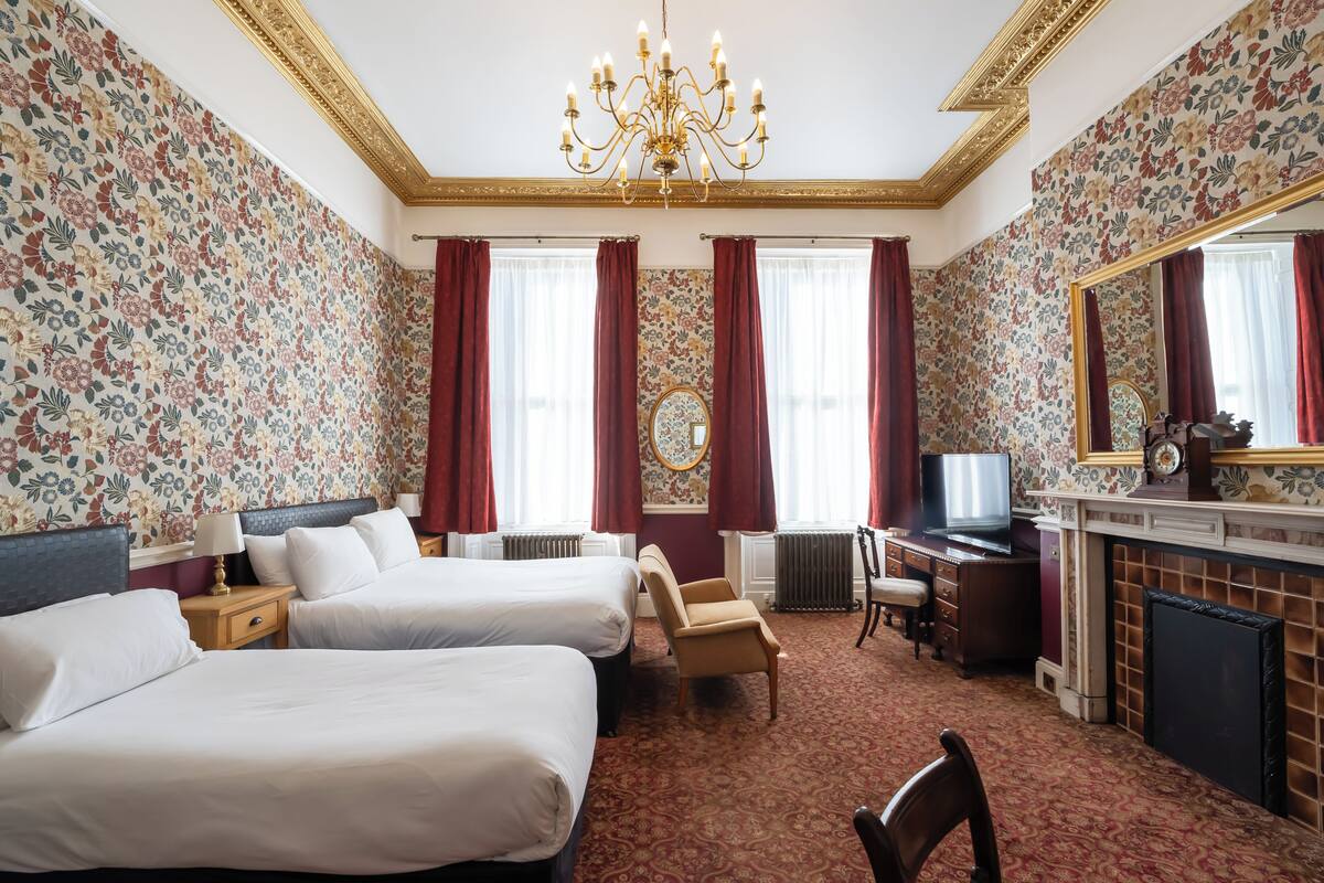 A hotel room with two beds, a desk, a television, a fireplace, and a large mirror.