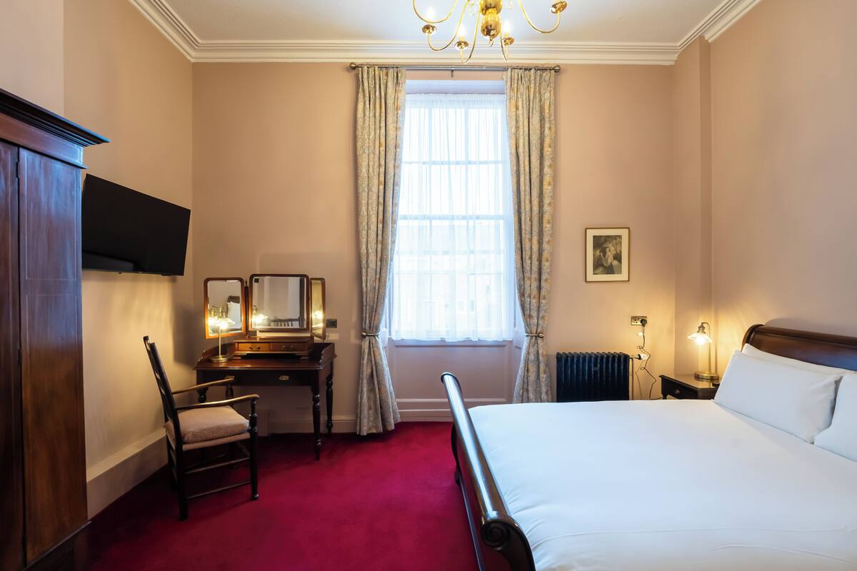 A hotel room with a bed, a desk with a chair, a window with curtains, a radiator, and a chandelier.