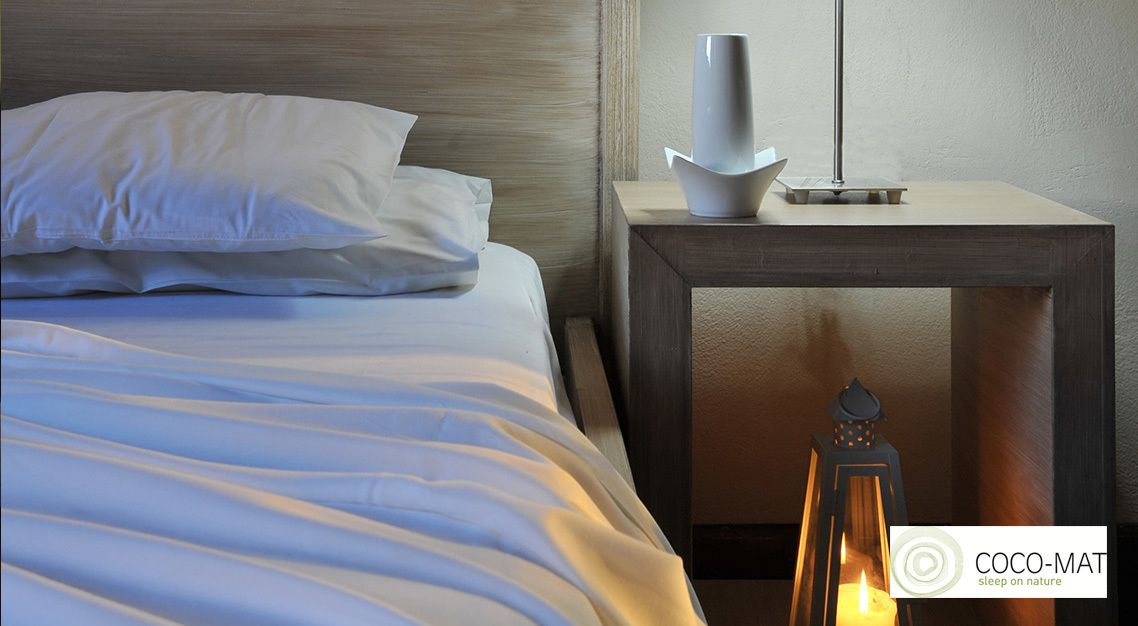 A neatly made bed with white linens and pillows, a bedside table with a modern lamp and a white vase, and a lit candle on the floor.