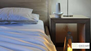 A neatly made bed with white linens and pillows, a bedside table with a modern lamp and a white vase, and a lit candle on the floor.