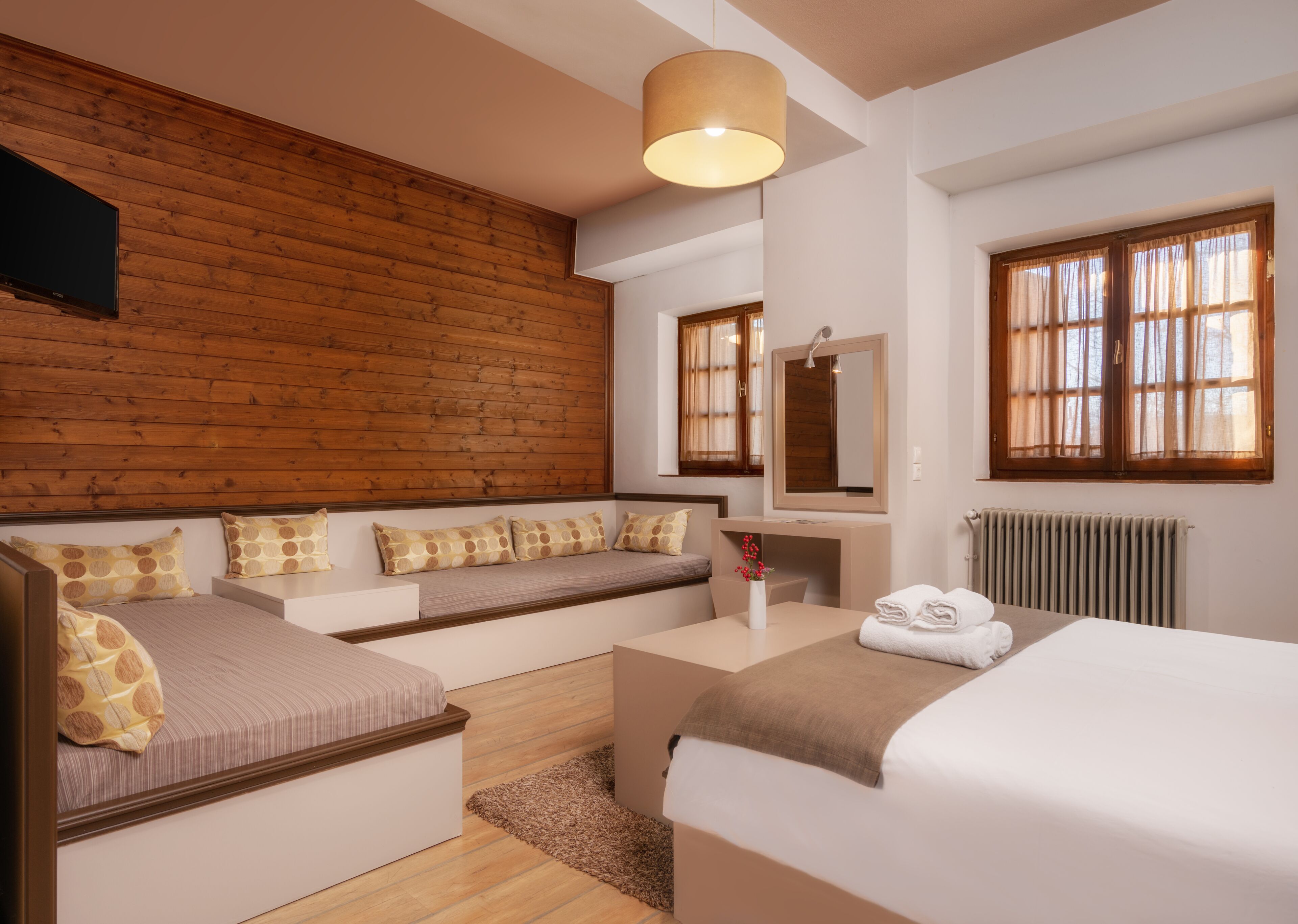A modern hotel room with a wooden accent wall, a sofa bed, a desk with a mirror, and a large window with curtains.