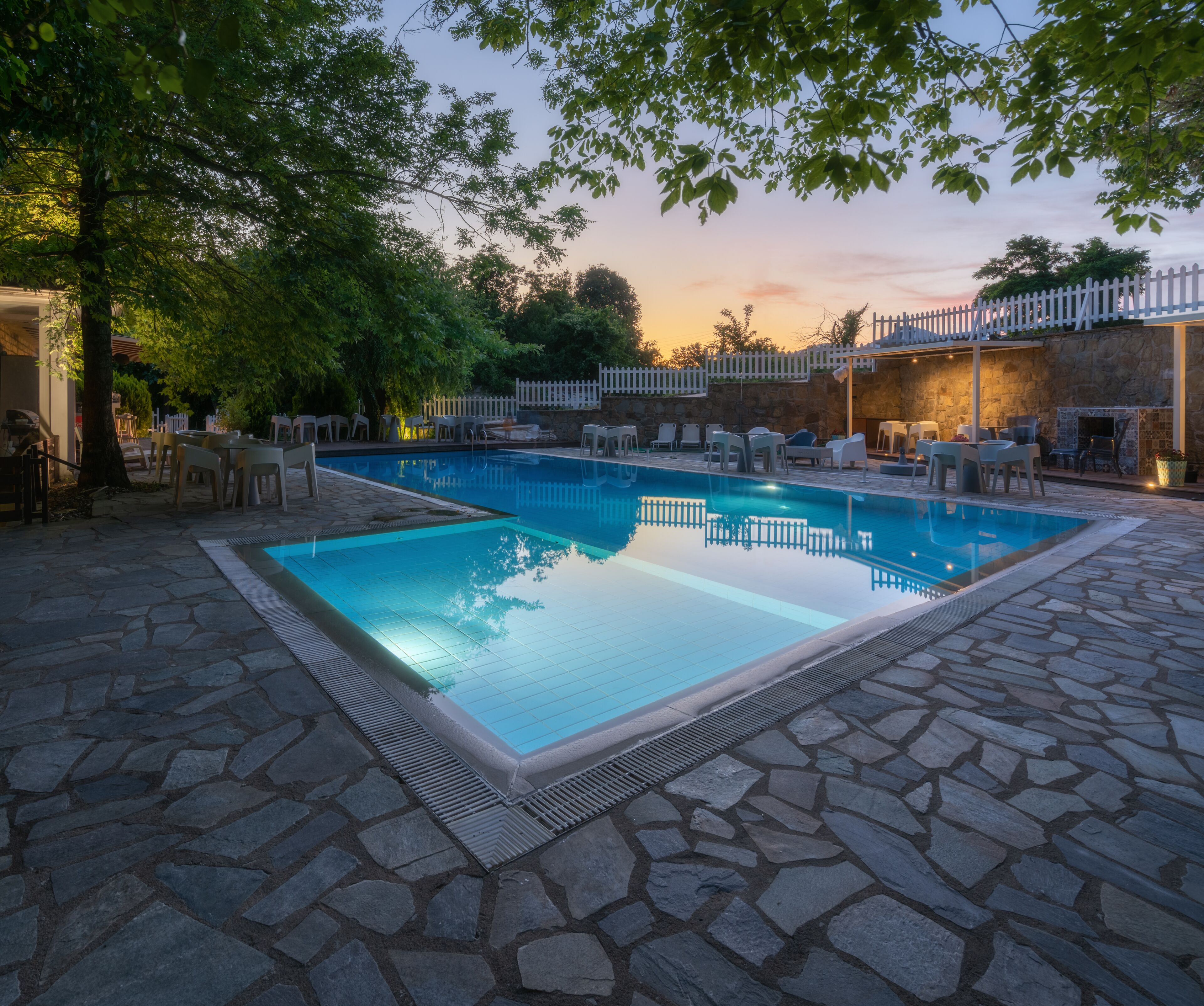 A rectangular swimming pool with a stone border, surrounded by lounge chairs and tables, set against a stone patio.
