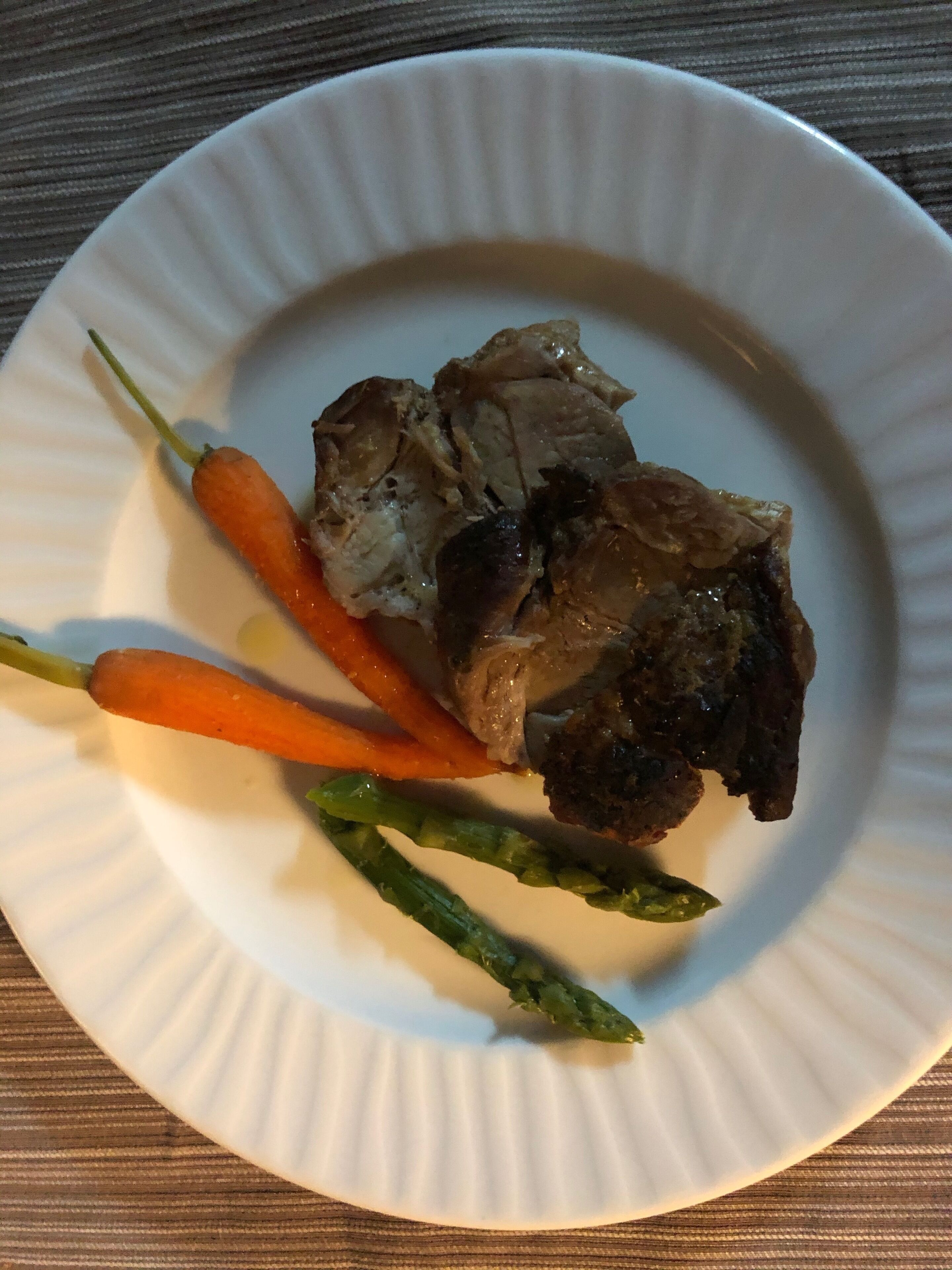 A white plate with a piece of meat, two carrots, and an asparagus on a wooden table.