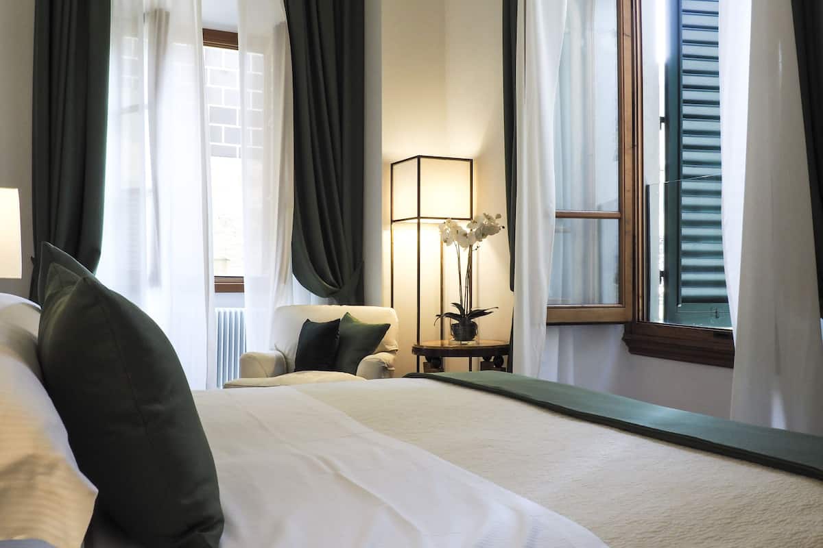 Quarto de hotel com uma cama grande, uma cadeira, uma mesa com abajur e uma janela com persianas.