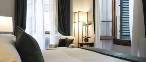 A hotel room with a large bed, a chair, a table with a lamp, and a window with shutters.