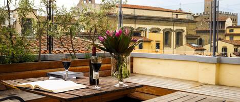 An outdoor seating area with wooden furniture, a table with a book and wine glasses, and a vase with flowers.