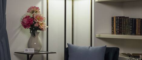 A modern room with a dark blue chair, a floor lamp, a vase with flowers, and a bookshelf with books.