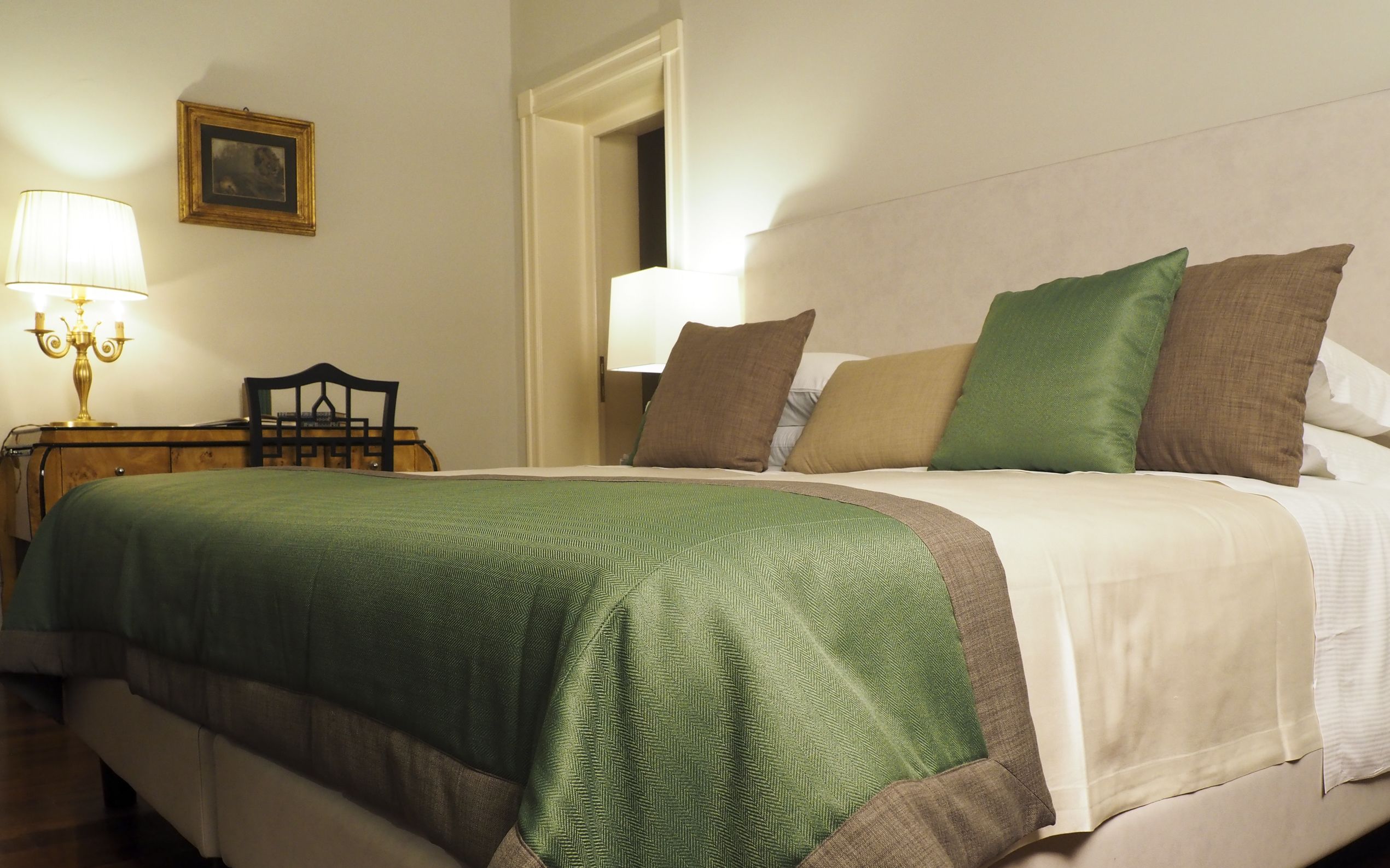 A neatly made bed with a green and beige bedspread, a wooden nightstand with a lamp, and a framed picture on the wall.